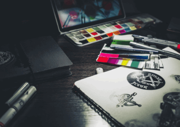 Edgy graphic design workspace with sketchbooks, color swatches, markers, and punk-style logo concepts under moody lighting