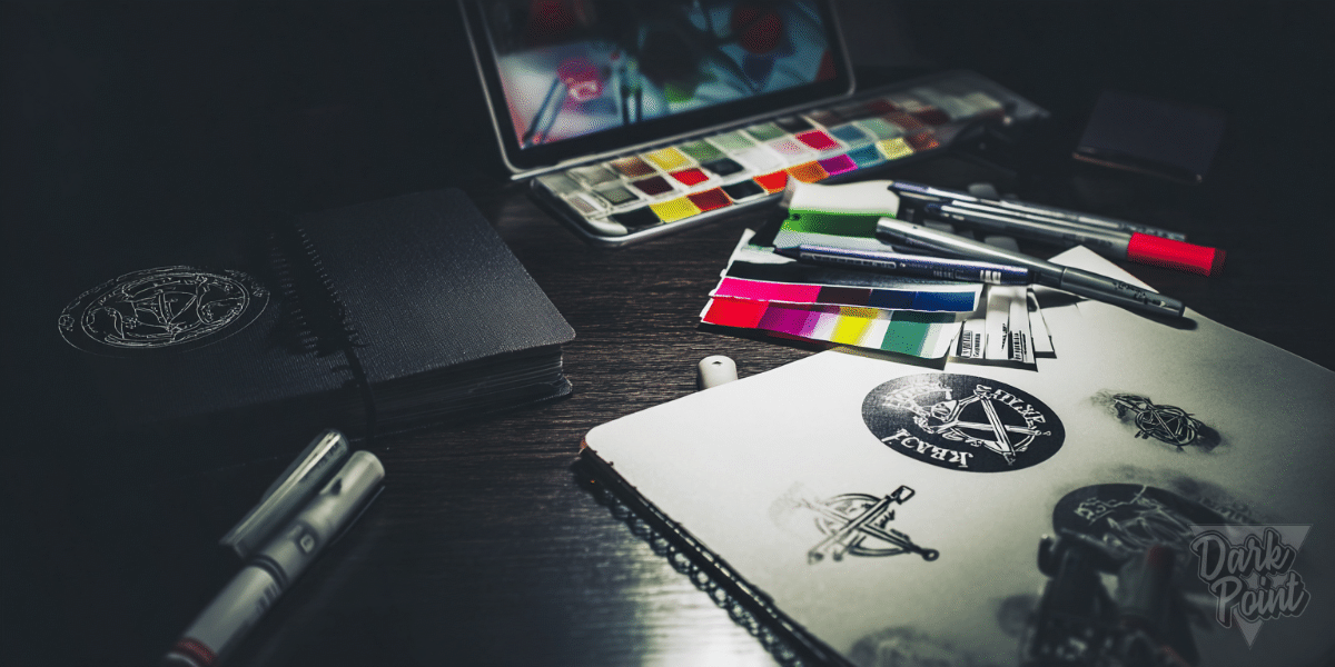 Edgy graphic design workspace with sketchbooks, color swatches, markers, and punk-style logo concepts under moody lighting